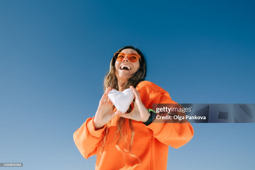 Cheerful woman in orange sweater and sunglasses smiles and holding heart-shaped snowball against blue sky