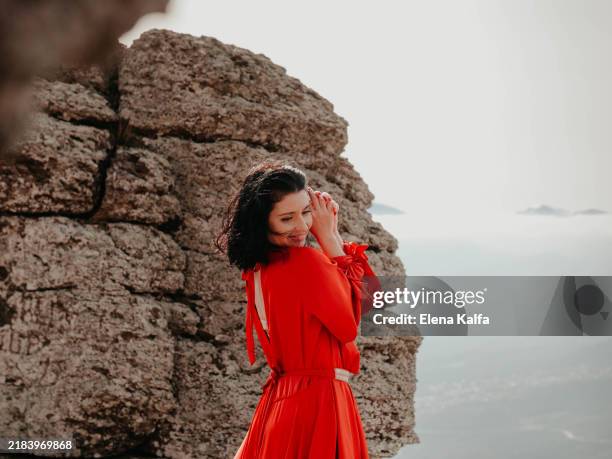 beautiful woman with dark hair in long red dress on mountain with view of clouds and sky. - red dress stock pictures, royalty-free photos & images