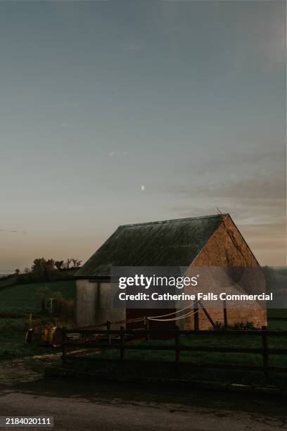 a rustic stone barn stands under the fading light of dusk with a crescent moon hovering above in the tranquil countryside - rustic barn stock pictures, royalty-free photos & images