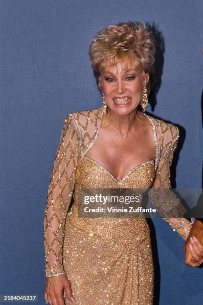American actress Barbara Eden, wearing a gold evening gown with gold detail sheer sleeves, against a blue background, attends a Friars Club of...