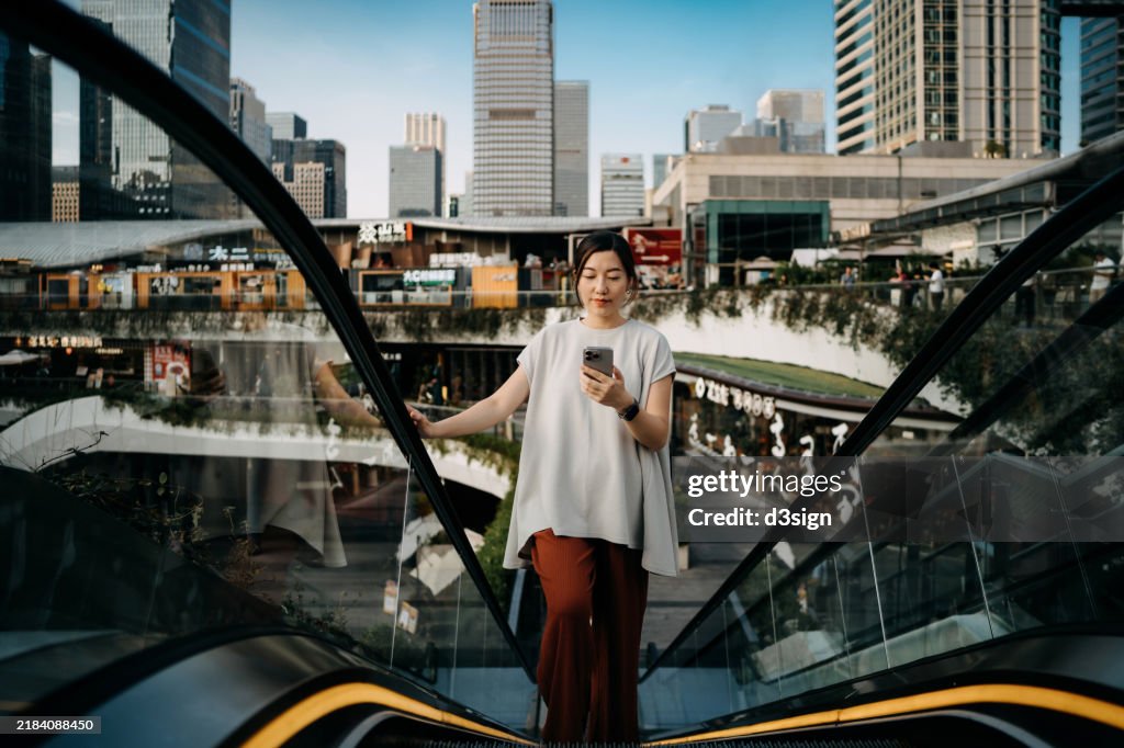 Young Asian businesswoman using smartphone while riding on escalator in a commercial complex in central business district, with highrise corporate buildings in the background. People, lifestyle and technology