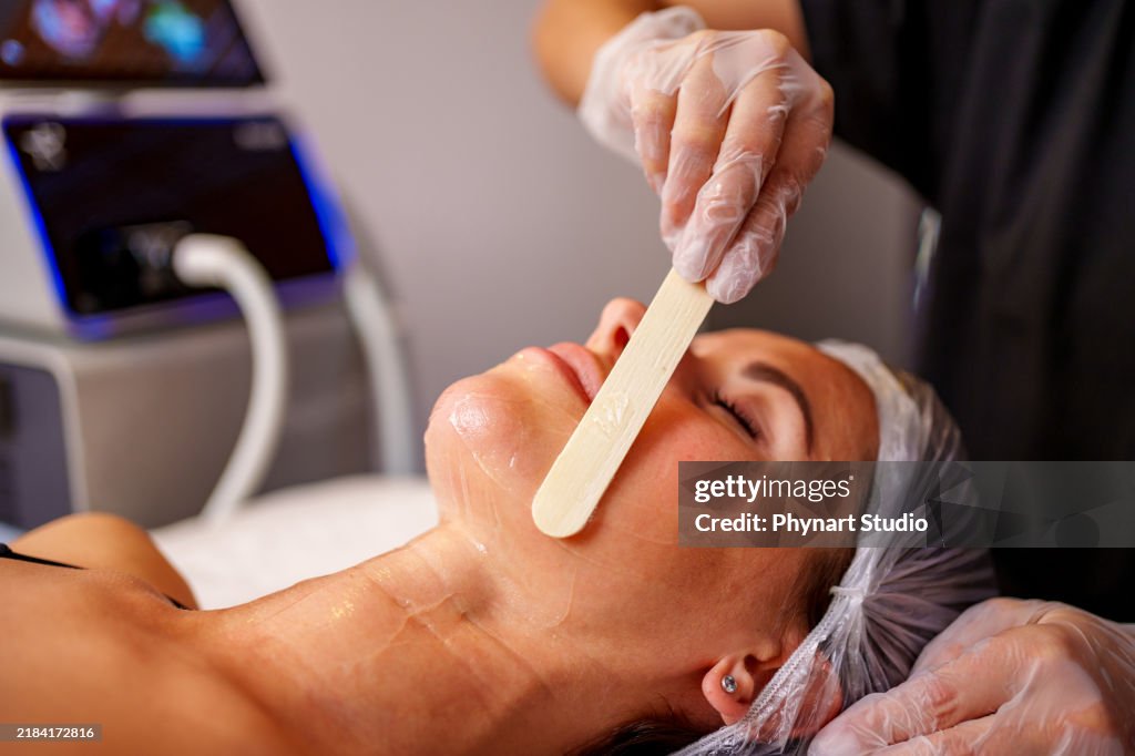 Closeup of beautician hands with wooden spatula applying cosmetic product gel peeling mask to beautiful young woman face