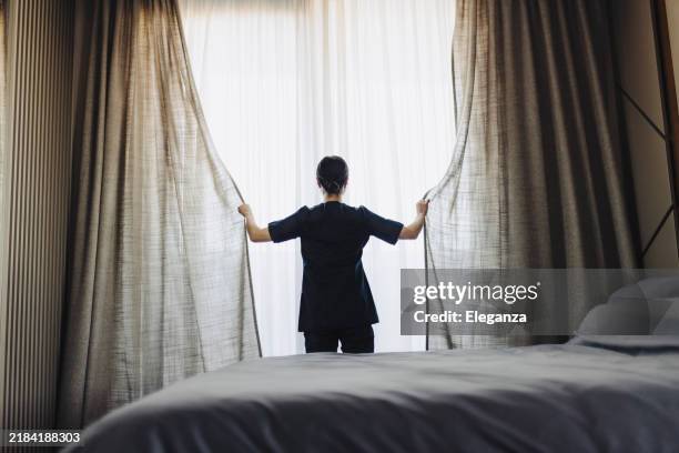rara vista de la camarera uniformada del hotel abriendo las cortinas blancas en la habitación del hotel - servicio de habitaciones fotografías e imágenes de stock