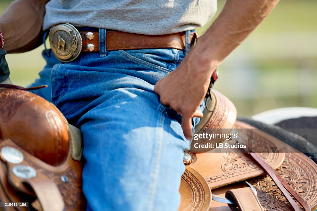 Close-up of a cowboy's midsection showing a silver belt buckle, blue jeans, and a detailed leather saddle