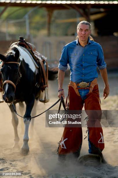 a confident man in cowboy attire walks beside a saddled horse in a dusty arena - calça de couro de cowboy imagens e fotografias de stock