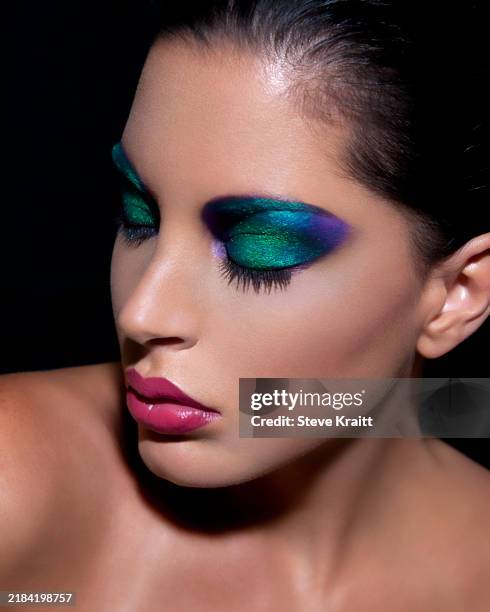 close-up of a woman with striking green and purple eye makeup and glossy pink lips against a dark background - eye make up stock pictures, royalty-free photos & images