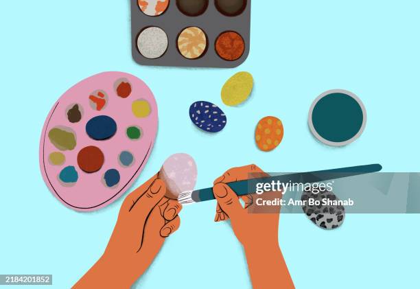 a pair of hands painting a colorful egg against a light blue background, with a palette and paintbrushes nearby - easter egg painting stock illustrations