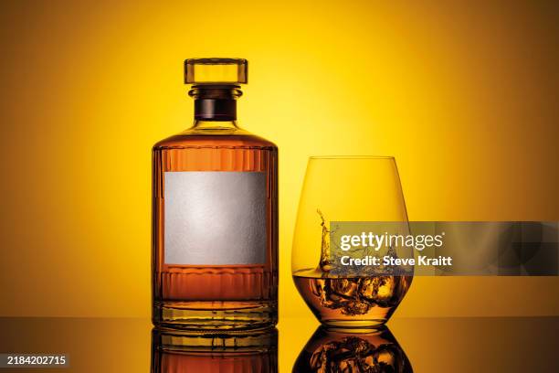 whiskey bottle and glass with a splash on a reflective surface against a yellow background - rubbing alcohol stock pictures, royalty-free photos & images