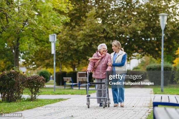 senior woman and her home caregiver spending a chilly, windy day outdoors in city park. autumn walk for elderly patient with walker. - osteoporose stock-fotos und bilder