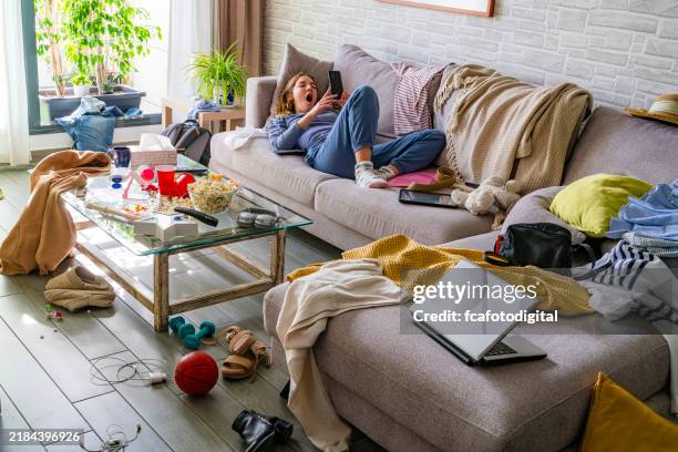lazy teenager using phone laying on the sofa of a messy living room - rommelig stockfoto's en -beelden
