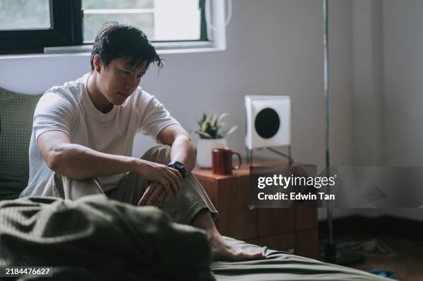 trauriger asiatischer chinese mann sitzt auf dem bett - winterdepression stock-fotos und bilder