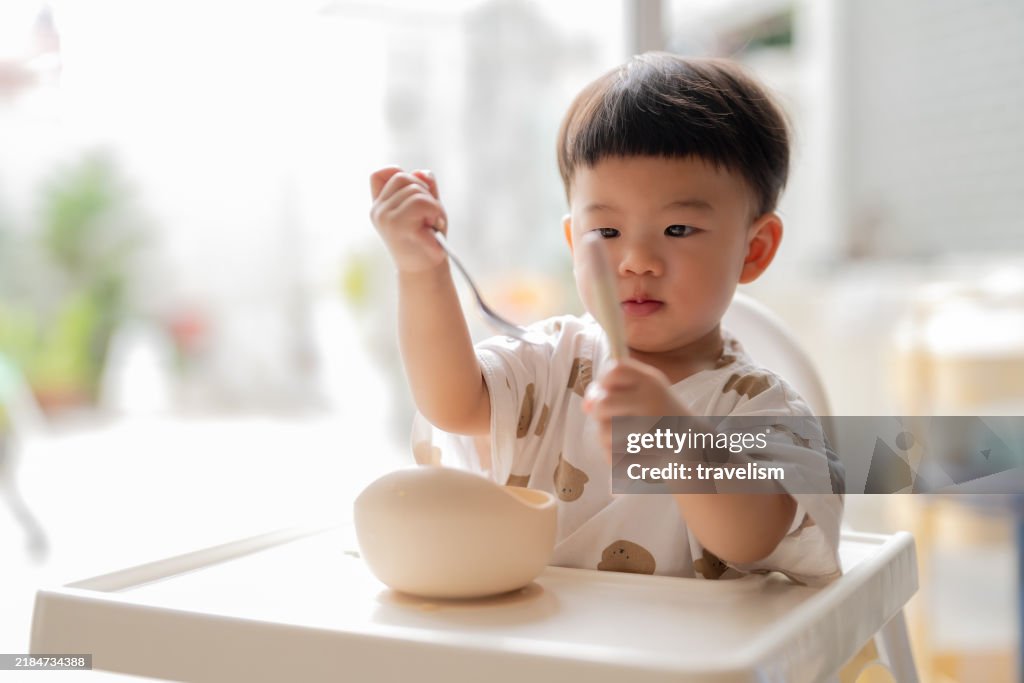 Adorable bebé asiático que come comida del plato por sí mismo mientras está sentado en casa de silla alta Hito del bebé, concepto de crecimiento y desarrollo