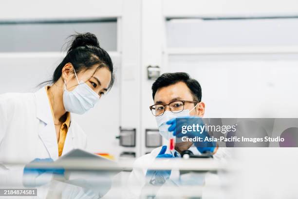 male and female scientists wearing protective face mask, looking through microscope and discussing over digital tablet in laboratory. - scientific research stock pictures, royalty-free photos & images