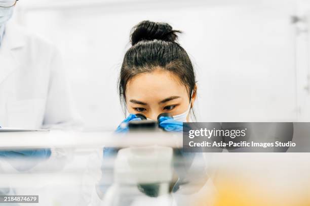 one chinese female scientist looking through microscope at work desk in lab. - wetenschappelijk onderzoek stockfoto's en -beelden