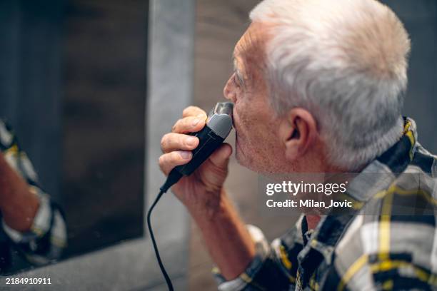 senior man shaves with an electric razor in bathroom - rasieren stock-fotos und bilder