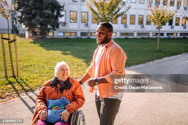 old people in wheelchair care outdoor by young adult volunteer talking and smiling - home carer stock pictures, royalty-free photos & images