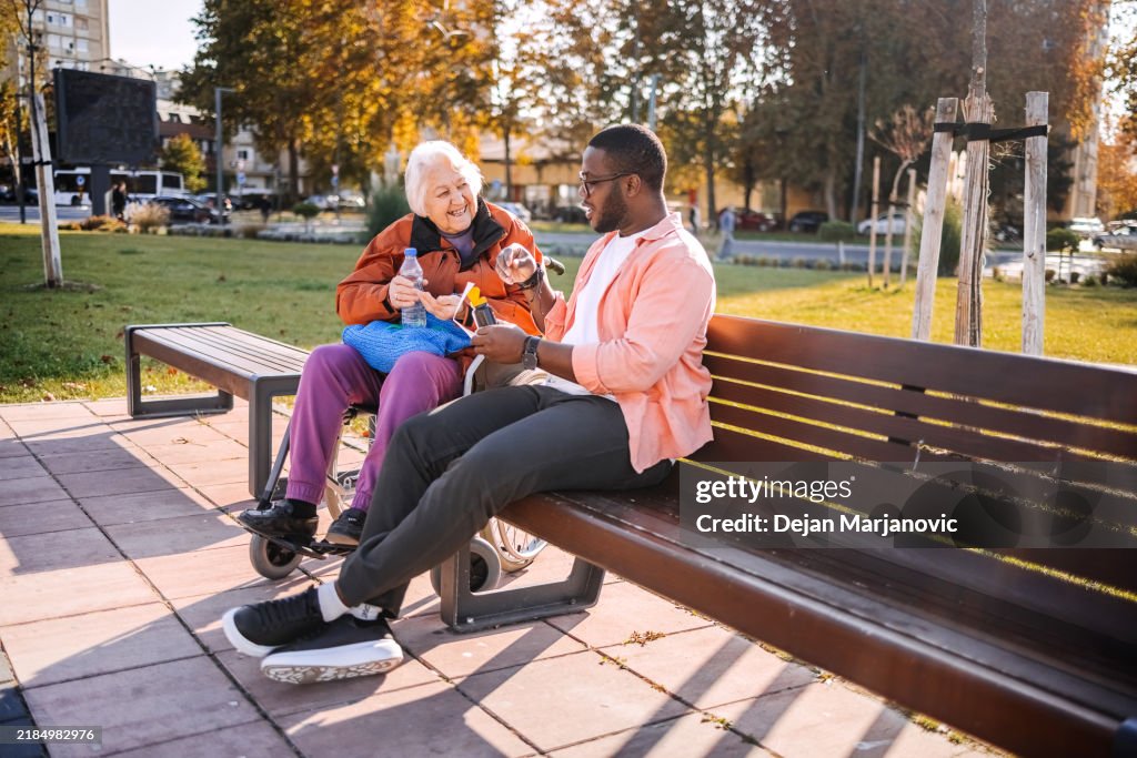 Old people in wheelchair care outdoor by young adult volunteer sitting on park bench and looking at prescription