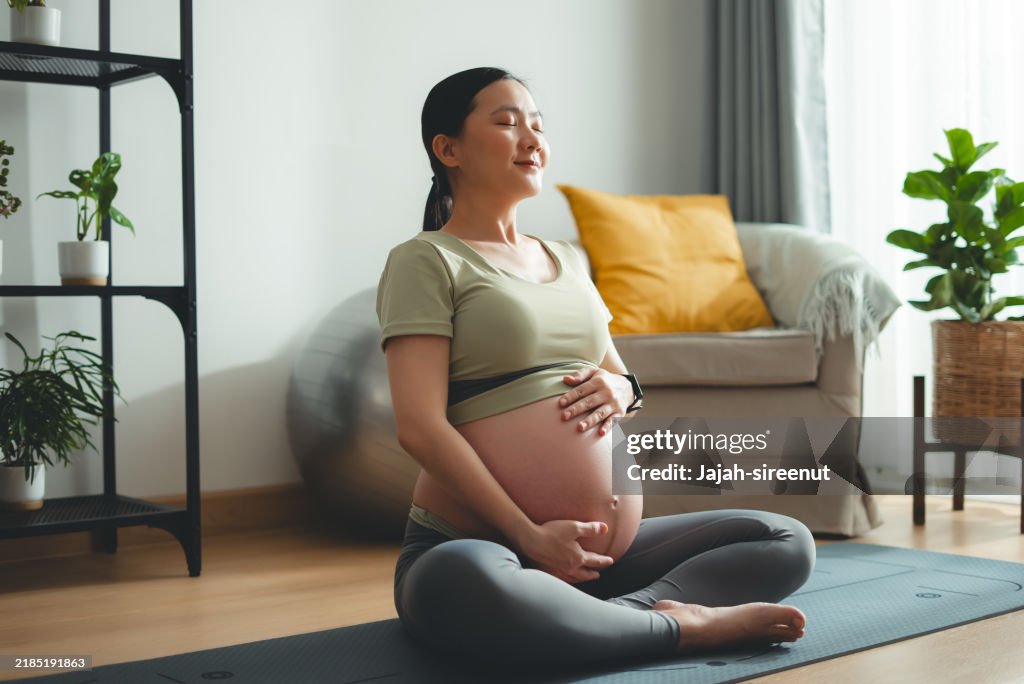 Asian pregnant woman practicing yoga on mat closed eyes do meditation in living room at home.