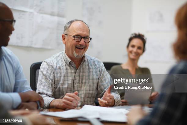 senior man talks smiles laughs in meeting with work colleagues while sitting at table in design office - real estate developer stock pictures, royalty-free photos & images