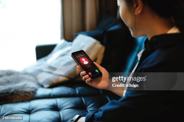 young asian woman receiving an incoming suspected call from unknown caller on her smartphone and rejecting the call at home. device screen showing warning sign as detected by network provider. phone scam. cyber security and fraud concept - broma pesada fotografías e imágenes de stock