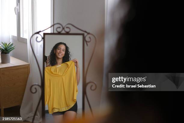 young woman trying on clothes and looking in mirror at home - vestirse fotografías e imágenes de stock