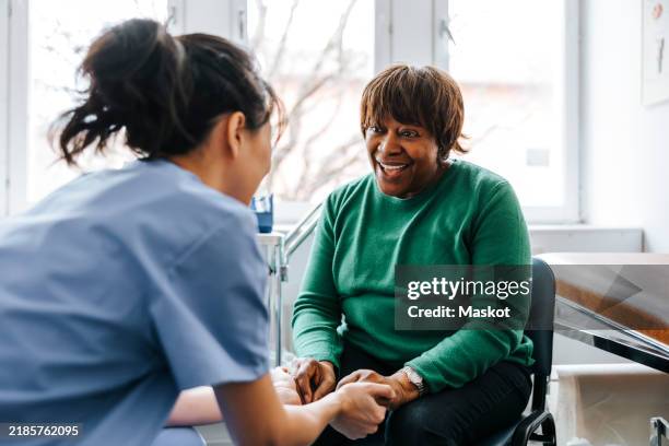 female healthcare worker holding hands of happy senior woman sitting in examination room at hospital - heilbehandlung stock-fotos und bilder