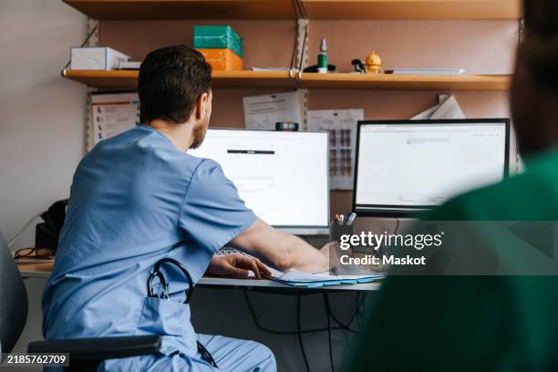 male physician in blue scrubs checking medical records on computer while sitting in examination room at hospital - medisch dossier stockfoto's en -beelden