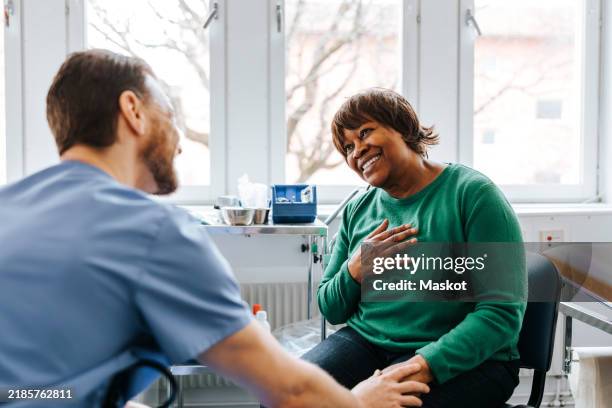 happy senior woman with hand on chest talking with male doctor while sitting in medical examination room - cuidados de saúde primários imagens e fotografias de stock