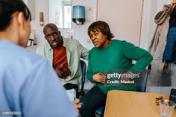 senior woman explaining stomach problems to doctor while sitting with son in examination room - stomachache stock pictures, royalty-free photos & images