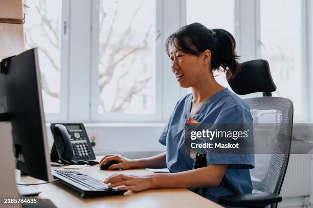smiling female healthcare worker working on computer while sitting in examination room at hospital - telephone switchboard stock pictures, royalty-free photos & images