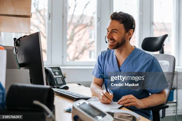 happy male medical professional writing while doing online consultation on video call through computer at clinic - doctor video call stock pictures, royalty-free photos & images