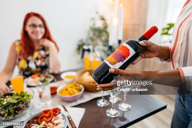 mid-adult woman serving a bottle of red wine to her female friend while having lunch in a domestic room - girls night in stock pictures, royalty-free photos & images