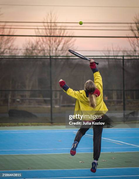 meistern des tennisaufschlags am herbstabend. zehnjähriges mädchen übt schlägertechniken mit gelber jacke auf blauem außenplatz - maschendrahtzaun stock-fotos und bilder