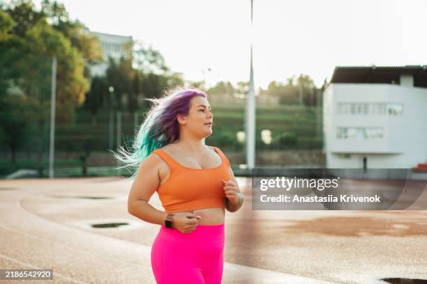 plus size woman with with pink and blue dyed hair in colorful sports wear running in morning on stadium. - tracksuit bottoms stock pictures, royalty-free photos & images