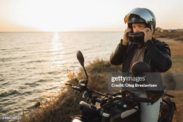 male biker adjusting helmet, sitting on motorcycle with sunset se view - motocicletta foto e immagini stock