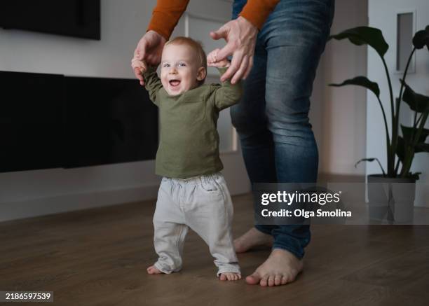 man teaching baby to walk. one year baby doing first steps with father support indoors. infant happiness, smiling , looking at camera - primeiros passos imagens e fotografias de stock
