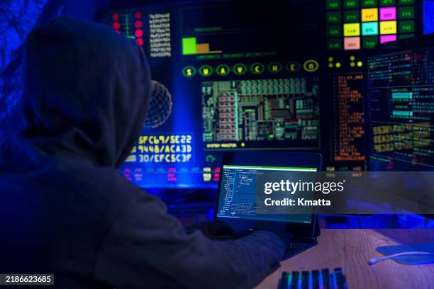 rear view of computer hacker in dark hall wearing hooded top hacks into a computer network, types software code on a keyboard and monitors a virus attack to hack into network systems and servers. - russian ethnicity stock pictures, royalty-free photos & images