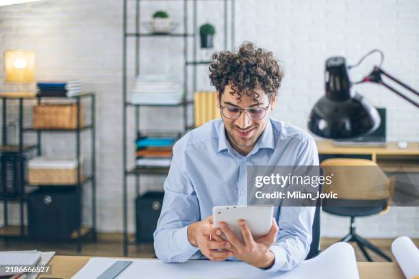 focused architect using tablet in bright modern office with blueprints on desk - software architect stock pictures, royalty-free photos & images