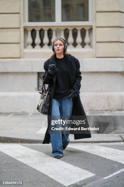Diane Batoukina wears Apple Airpods Max, a black long coat from Zara, a black cashmere pullover from Massimo Dutti, blue denim wide leg jeans from...