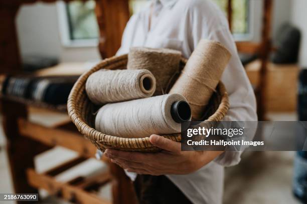 woman holding basket full of threads, standing next to wooden loom. small weaving business making handmade products from upcycled materials. - macchina per cucire foto e immagini stock