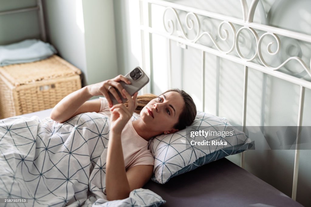 Junge Frau, die morgens auf dem Bett liegt und auf ihr Handy schaut