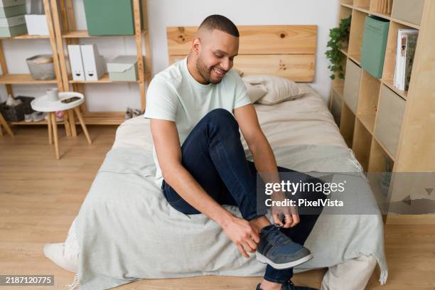 young man tying his shoes while sitting on bed in his bedroom - tying shoelace stock pictures, royalty-free photos & images