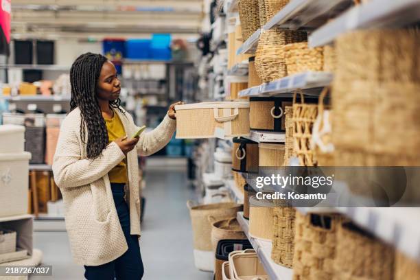 une femme noire joyeuse choisit le panier de rangement qu’elle achète dans un grand magasin et se prépare à les prendre en photo avec son téléphone portable, afin de pouvoir décider plus tard lequel elle préfère - équipement domestique photos et images de collection