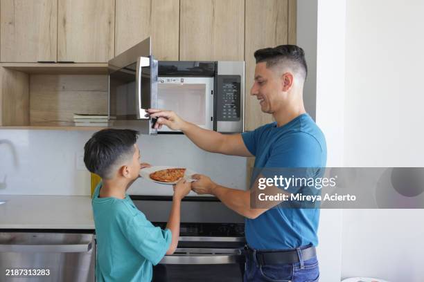 a latinx father helps his son warm up a slice of pizza in the microwave while wearing casual clothing. - microwave stock pictures, royalty-free photos & images