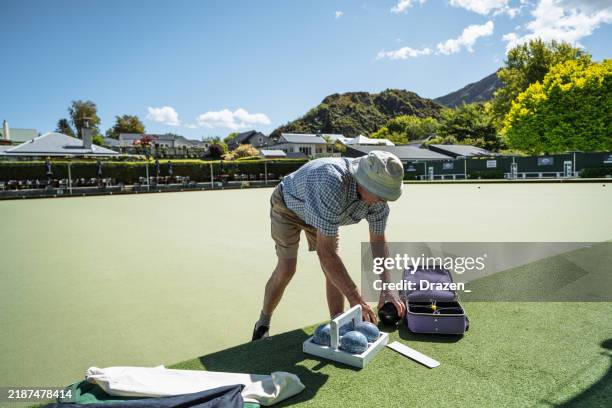 senior man playing lawn bowls on a sunny day at an outdoor bowling club, wearing casual summer clothing and a sunhat. man is packing his bowls in the bag after the game. - demography stock pictures, royalty-free photos & images