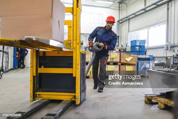 focused adult mixed male warehouse worker with a helmet lifting a big cardboard box with a pallet truck in a big warehouse - pallet truck stock pictures, royalty-free photos & images