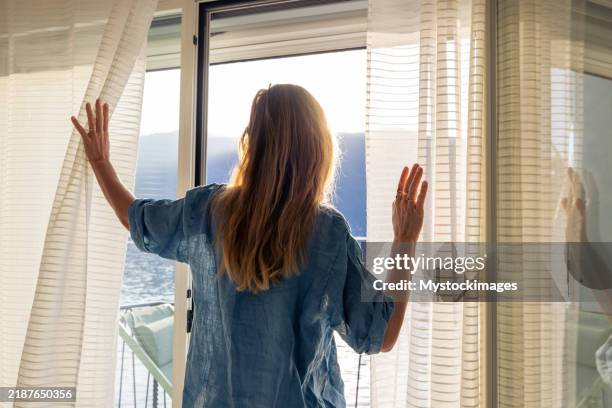 woman opening curtains in a sunlit room overlooking the sea - comfort zone stock pictures, royalty-free photos & images
