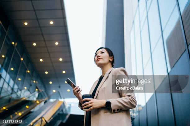 professional young asian businesswoman using smartphone while taking a coffee break, standing against modern financial skyscrapers in an urban setting. looking positive and motivated. professional occupation. business, finance and investment concept - azië en pacifisch gebied stockfoto's en -beelden