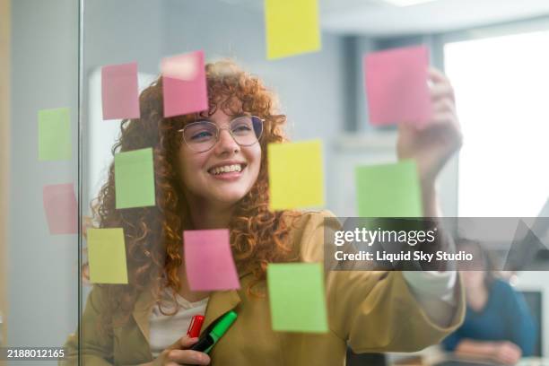 young businesswoman planning strategy using sticky notes on glass wall in office - papel adesivo imagens e fotografias de stock