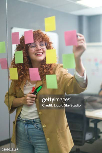 junge geschäftsfrau plant und brainstormt mit haftnotizen an glaswand im büro - projektmanagement stock-fotos und bilder